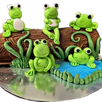 Log-shaped cake textured like bark, with five green fondant frogs, vines and a small blue pond on a silver board