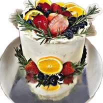 Tall white-iced cake on a silver platter, topped with strawberries, blueberries, orange slices and sprigs of rosemary