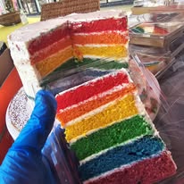 Tall white-iced cake with a slice held up in a blue glove, showing six coloured sponge layers from red through to purple