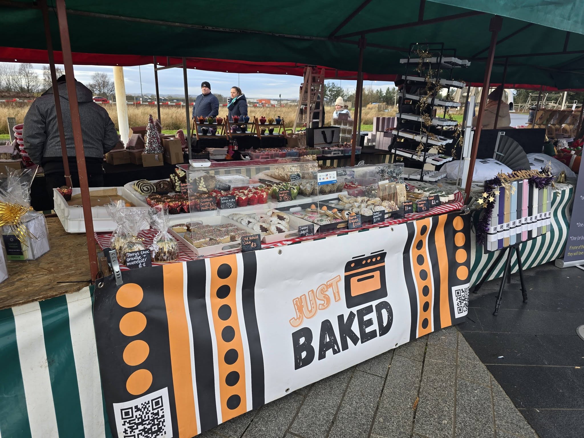 Just BAKED stall under a green awning, with the orange and black banner across the front and homemade cakes set out on the table